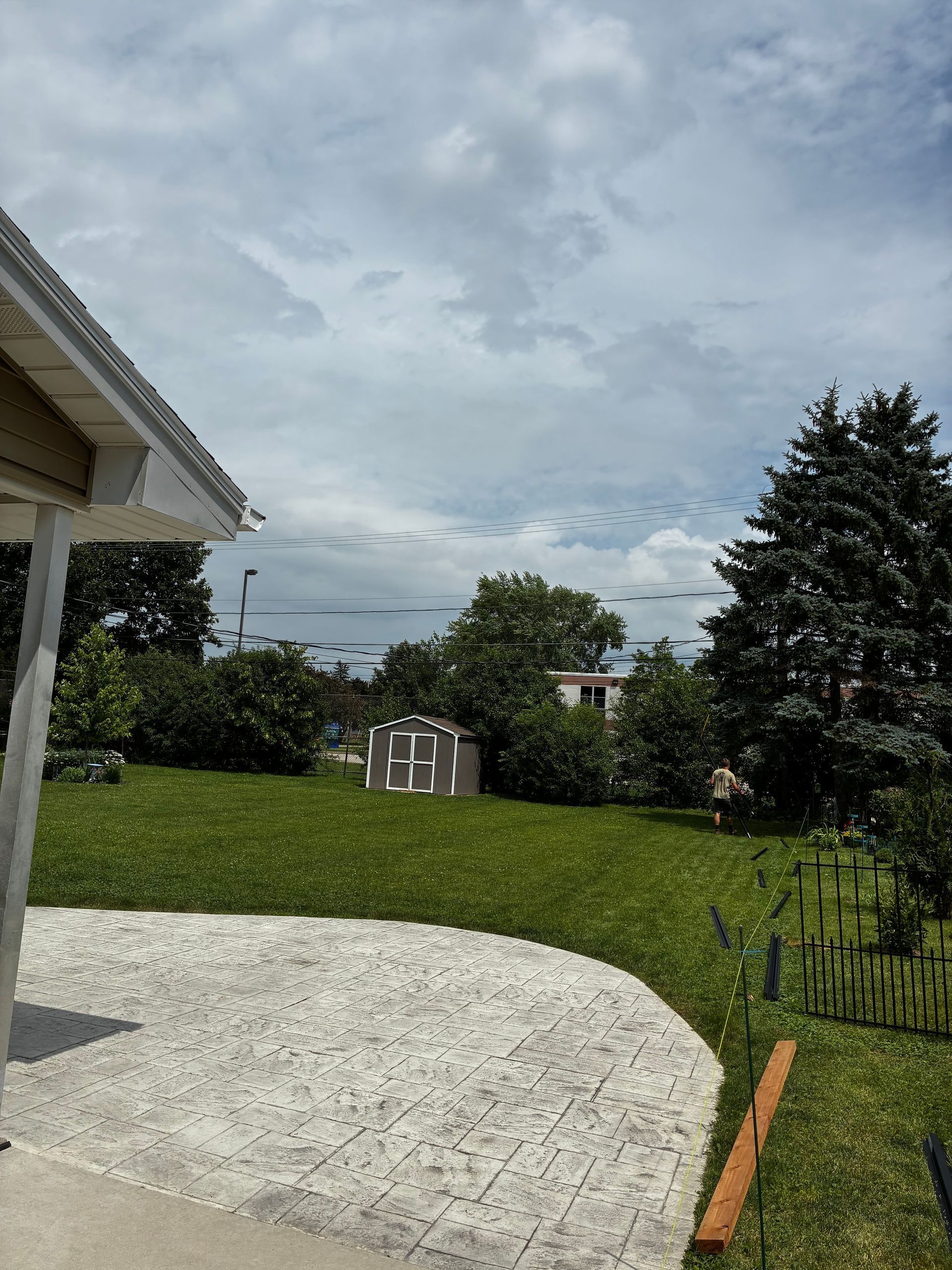 Backyard with curved stone patio, green lawn, trees, and cloudy sky beside a white house