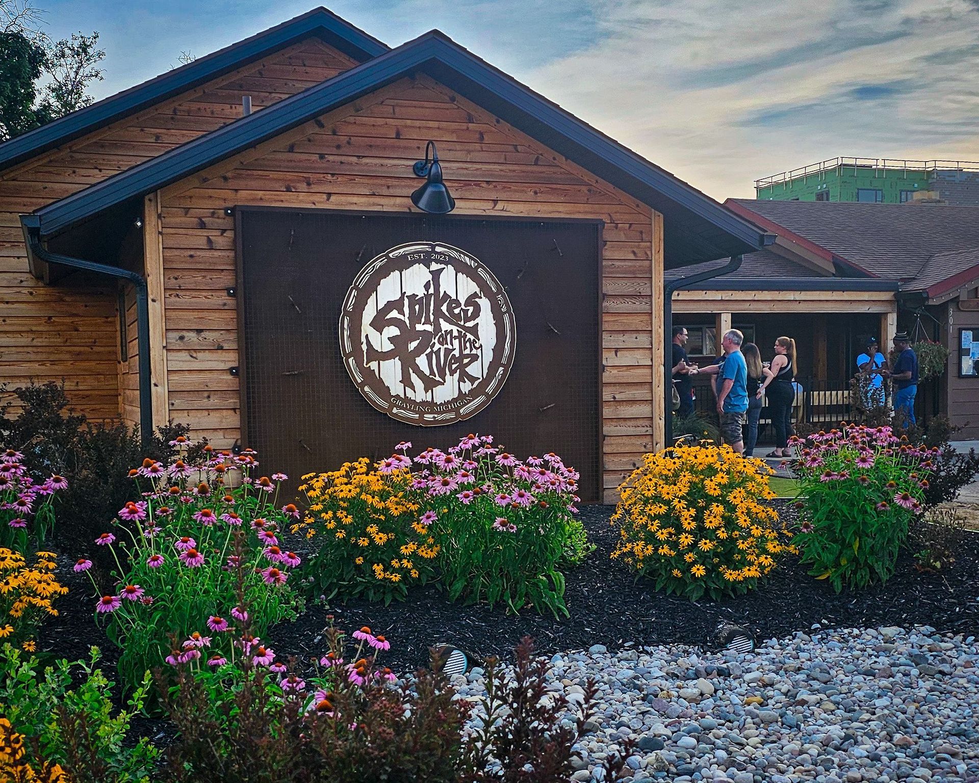 Rustic wooden building with circular sign, colorful flowers, and people gathered by the entrance at dusk