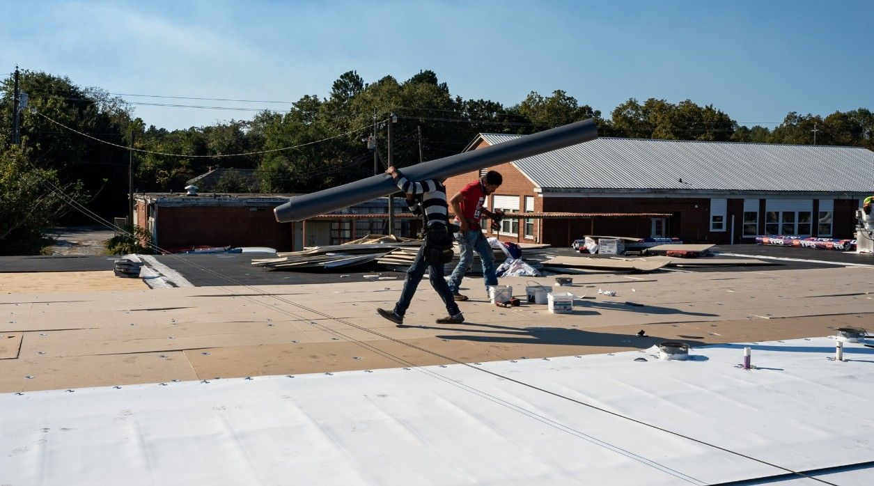 Two construction workers carrying a long gray beam across a rooftop on a sunny day.