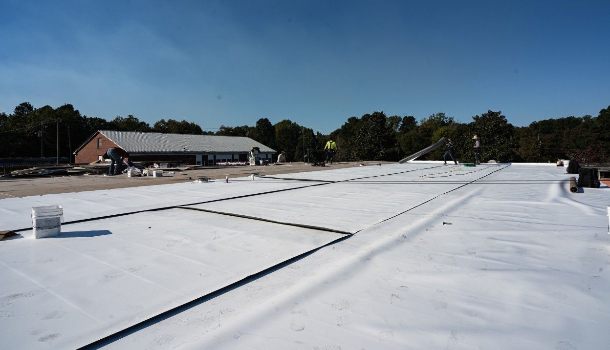Flat white roof under construction; building, trees, and clear sky in the background.