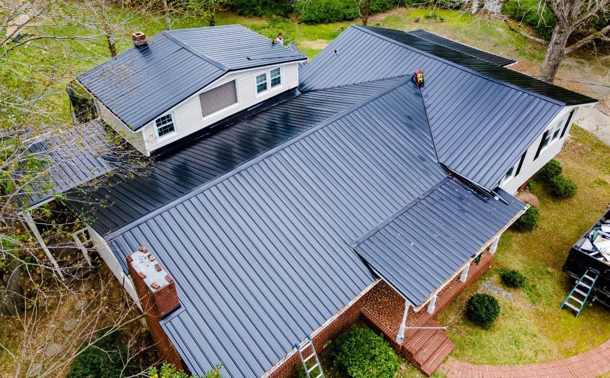 Black metal roof on a two-story house with a chimney, red brick, and green landscaping.