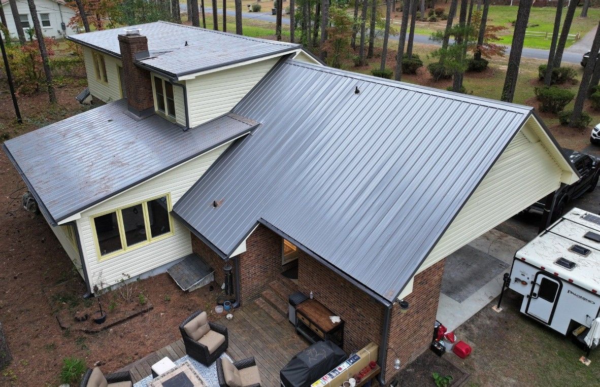 An aerial view of a two-story house with a dark metal roof. The house is surrounded by trees.
