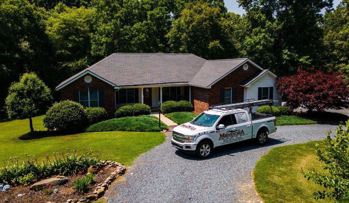 A white pickup truck parked in front of a brick house with a gray roof. Lush green trees surround the house.