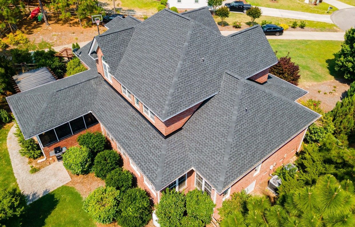 Aerial view of a brick house with a dark gray shingled roof surrounded by green trees and grass.