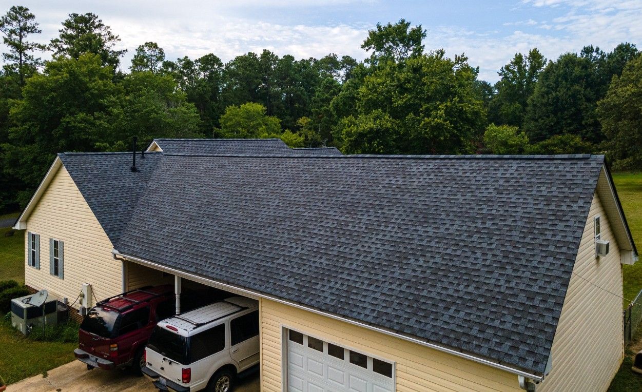 Yellow house with dark grey shingle roof, a carport with two vehicles, and a closed garage door. Trees in the background.