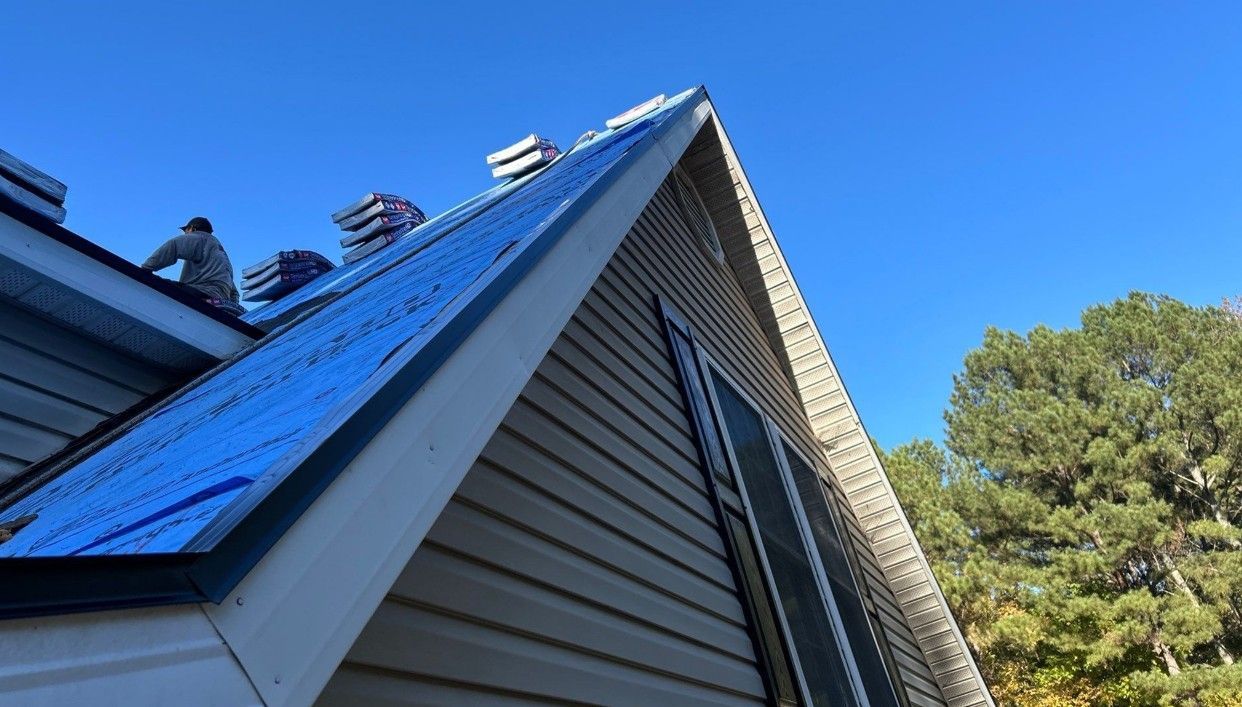 Roofer on a gable roof, blue underlayment visible, stacks of shingles, clear blue sky, sunny.
