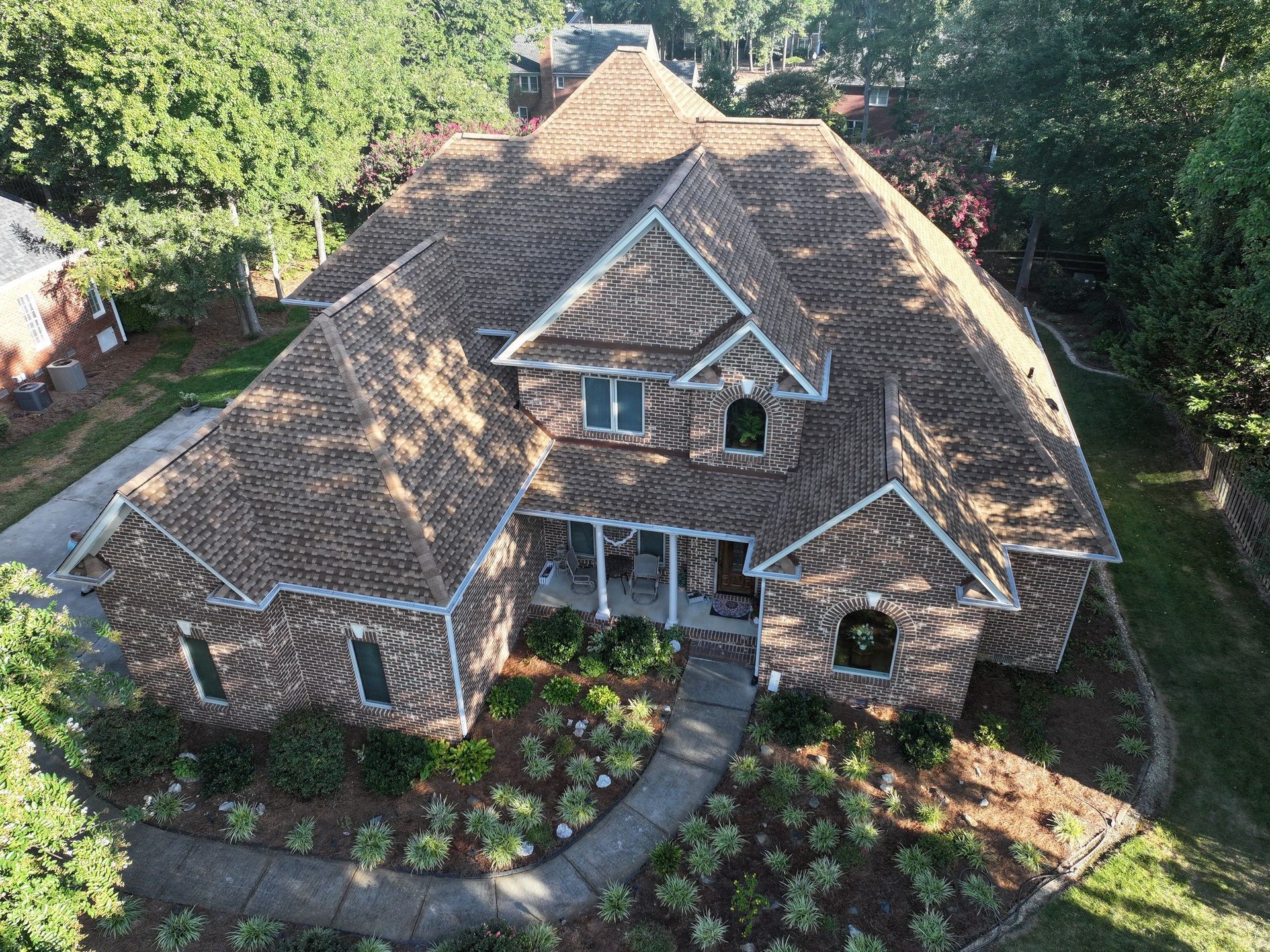 Aerial view of a brown brick house with a complex brown roof and manicured landscaping.