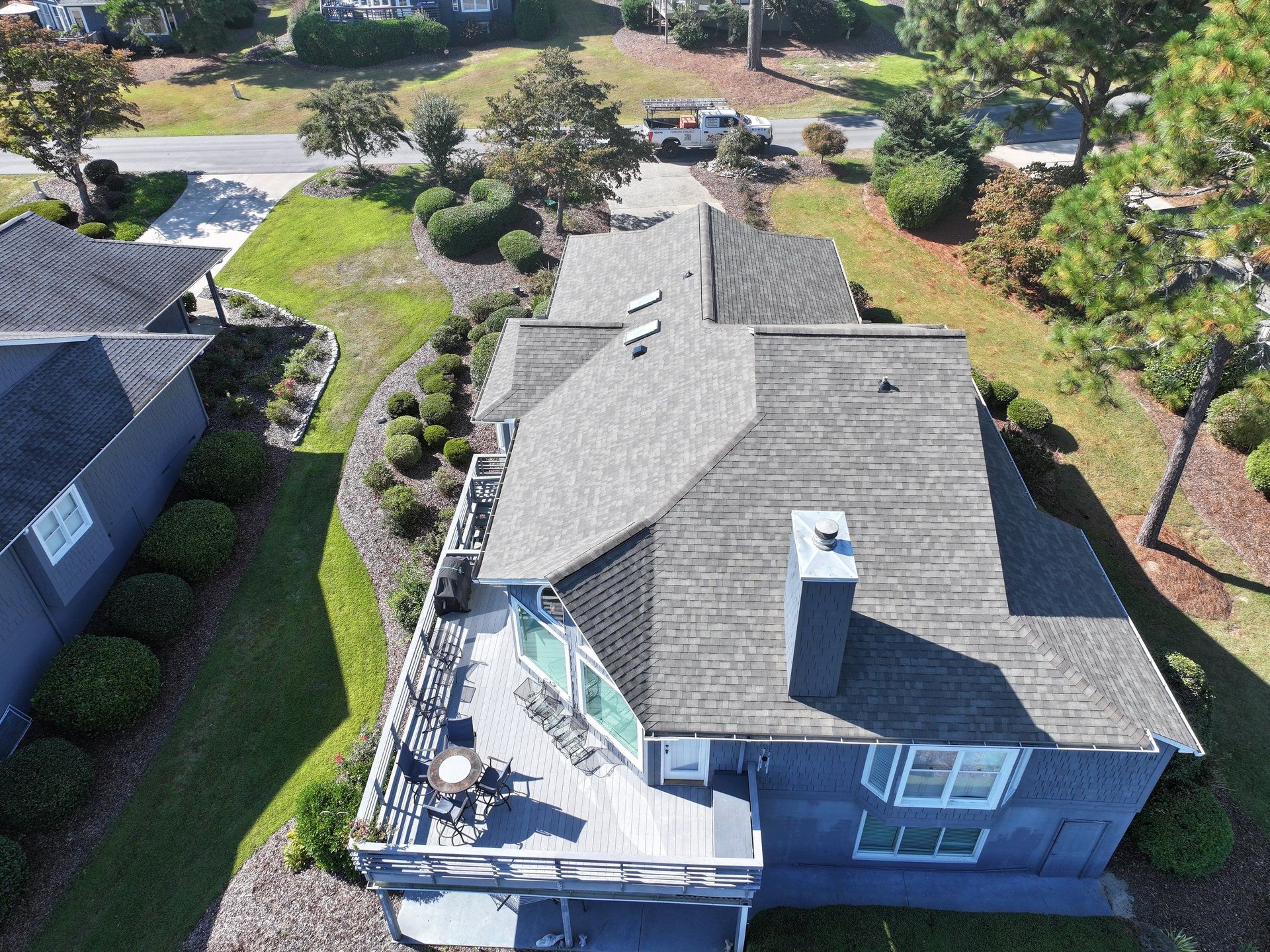 Aerial view of a gray house with a deck, dark roof, and lush green landscaping.