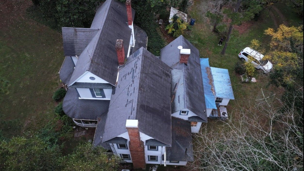 Overhead view of a multi-gabled house with a dark roof surrounded by trees and overgrown yard.