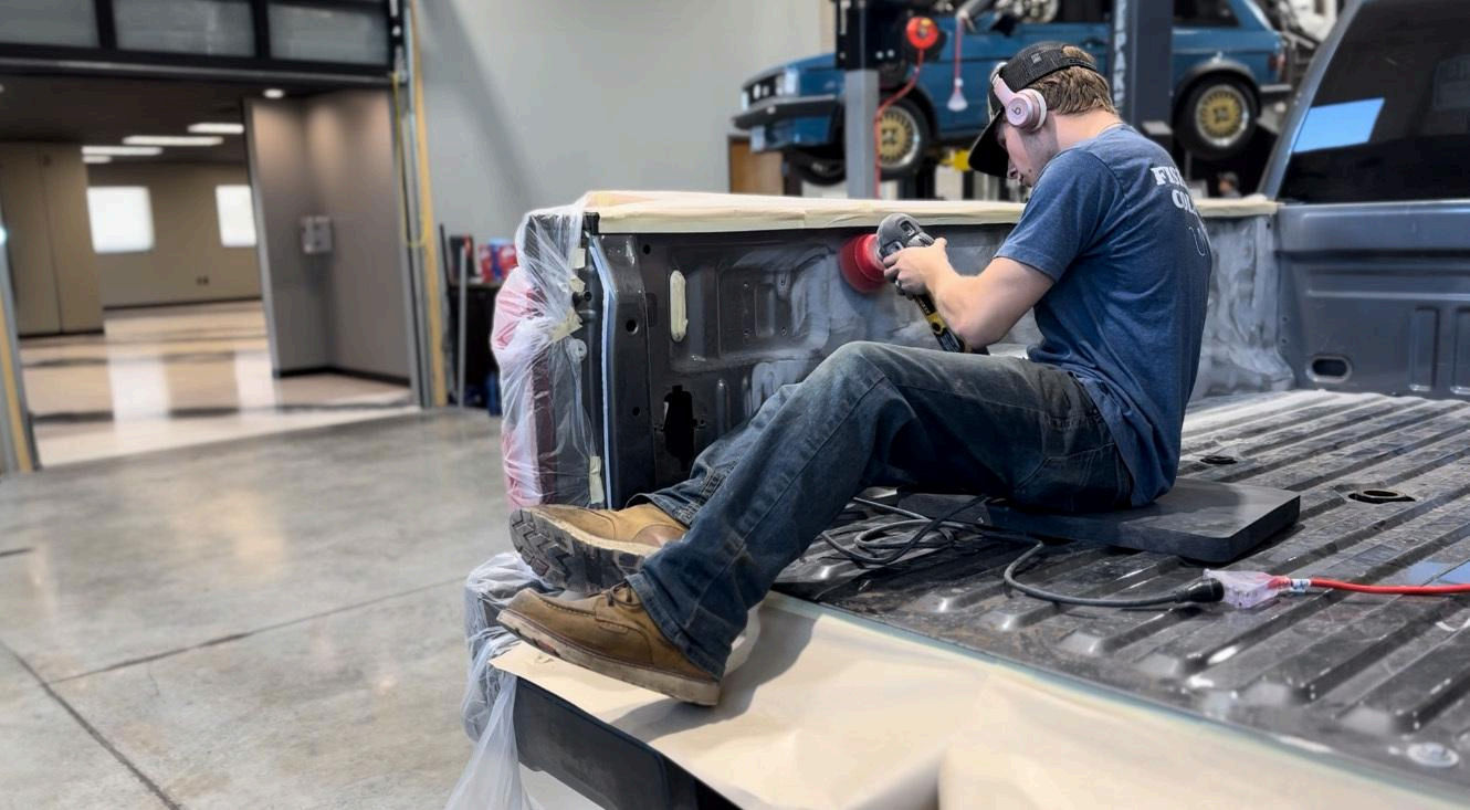 A person sitting inside a truck bed uses a power sander on the side panel inside an automotive workshop.