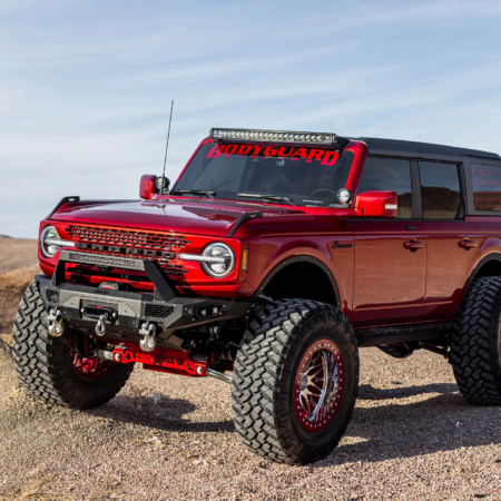 A modified red Ford Bronco with large off-road tires, a lifted suspension, and a front bumper, parked on a gravel surface.