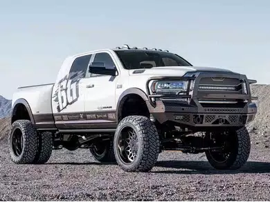 Modified white and brown RAM dually pickup truck with large off-road tires and a custom steel bumper on rocky terrain.