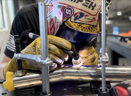 A welder wearing a sticker-covered mask uses a TIG torch to join metal pipes held by clamps on a workbench.