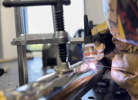 A gloved welder uses a TIG torch to fuse a metal pipe, secured by a C-clamp on a workbench.