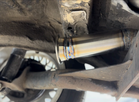 A freshly welded stainless steel exhaust pipe section mounted under the chassis of a vehicle near a tire and suspension.
