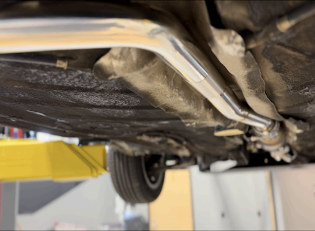 A low-angle view of a car’s undercarriage on a yellow lift, focusing on a newly installed, shiny metal exhaust pipe.