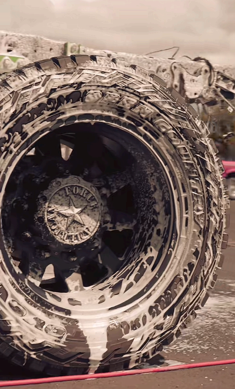 A large, black off-road truck tire and wheel covered in white soap suds during a car wash.