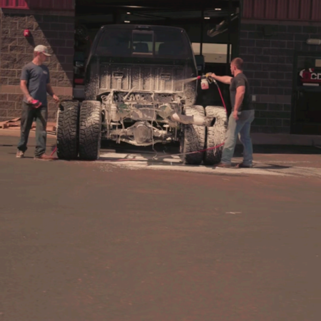 Two people work outside a shop cleaning the exposed undercarriage and dually rear tires of a truck with a power sprayer.