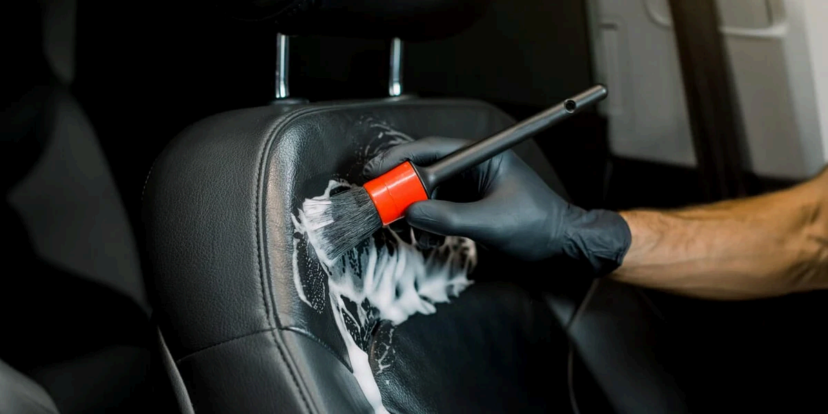A person wearing black gloves uses a soft-bristled brush to scrub white cleaning foam into a black leather car seat.