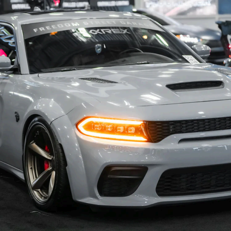 A grey widebody Dodge Charger with bronze wheels, LED daytime running lights, and a windshield banner at an indoor event.