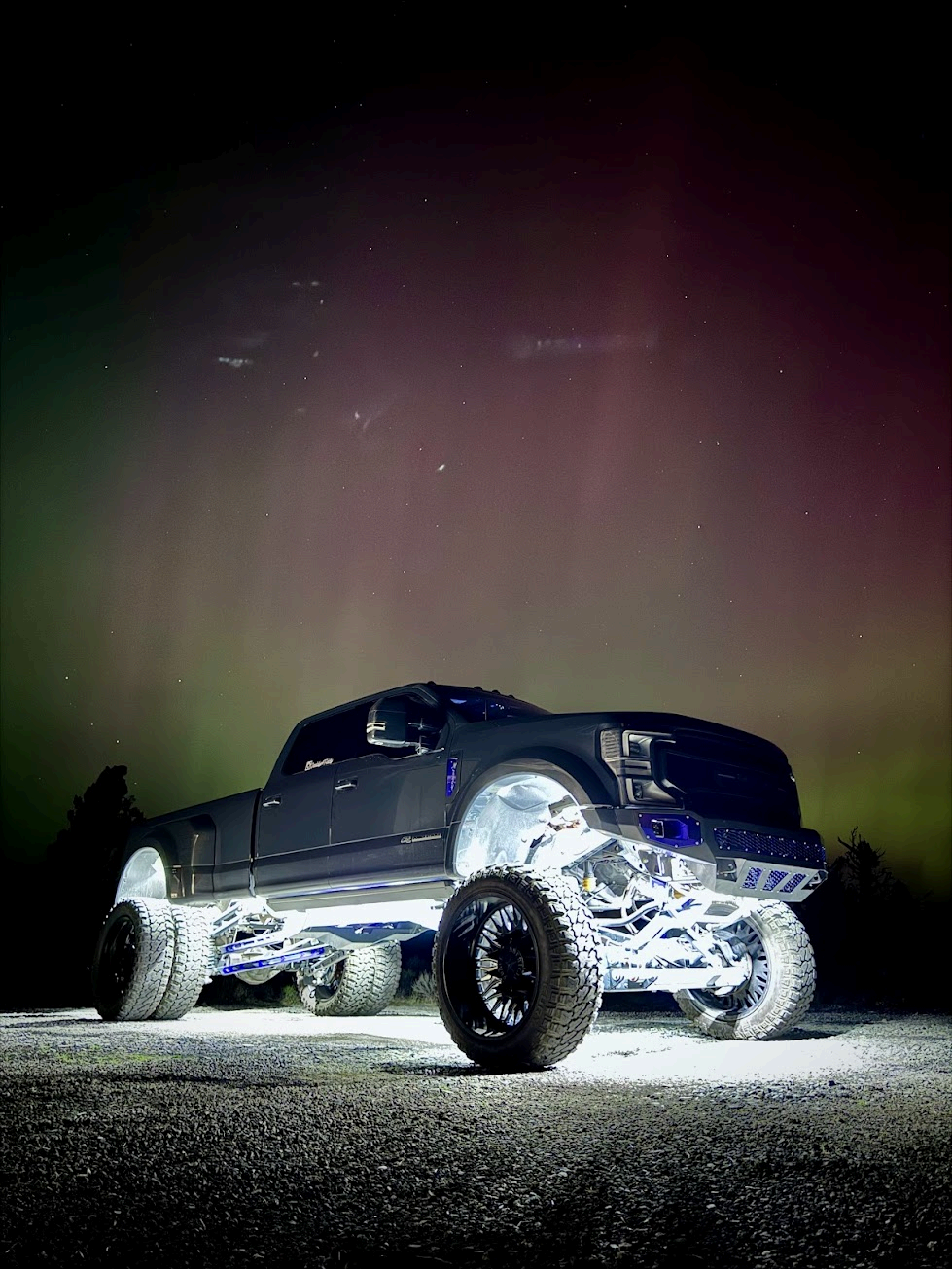 A lifted, dark-colored pickup truck with bright white underglow lights parked at night under a vibrant green aurora.