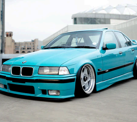 A lowered, bright turquoise BMW E3 Series sedan parked on concrete in front of an architectural structure.