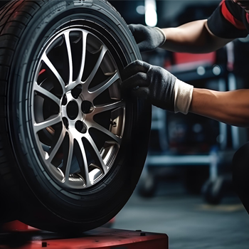 A mechanic wearing gloves carefully handles a car tire and silver rim in a workshop setting.