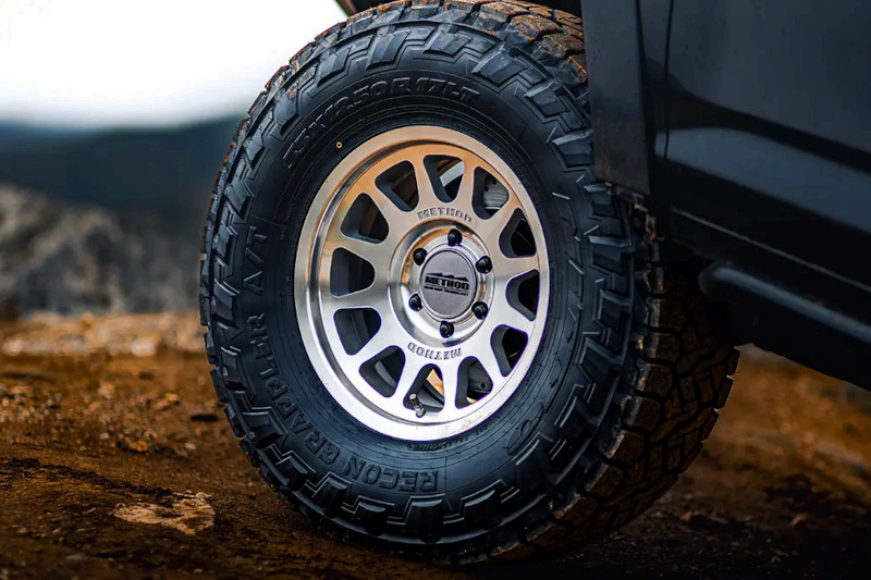 A close-up shot of a rugged, off-road tire mounted on a silver alloy wheel, parked on a rocky, dirt-filled terrain.