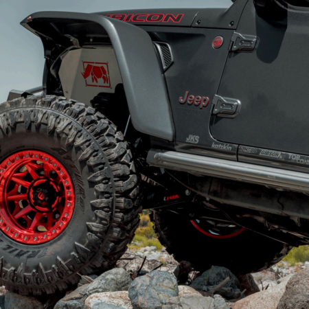 A close-up shot of a dark Jeep Rubicon with red wheels and branded fender liners navigating rugged, rocky terrain.