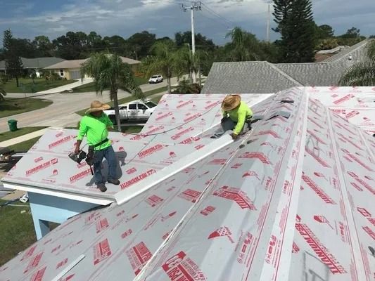 Two roofers in green shirts and straw hats working on a roof with a white underlayment in a suburban setting.
