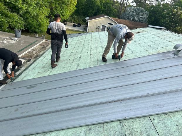 Three workers installing metal roof panels on a building roof, sunny day.