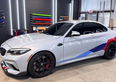 Silver BMW M2 with M-colored stripes, black rims, parked indoors under bright lights.