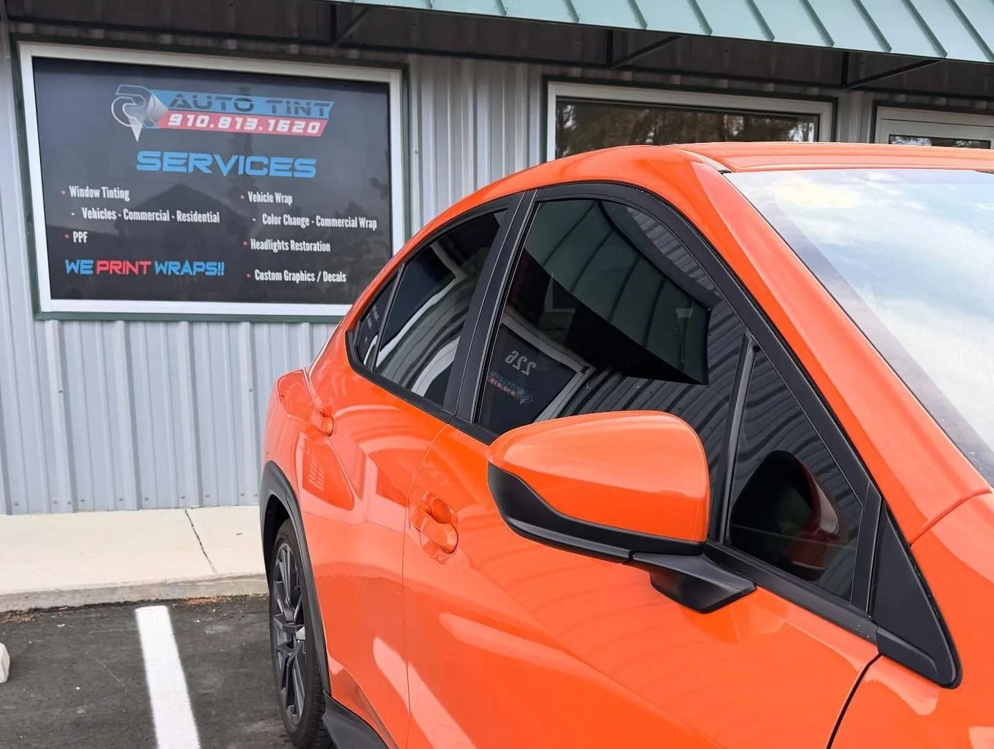 Orange car parked outside a business with a sign advertising services including window tinting and wraps.