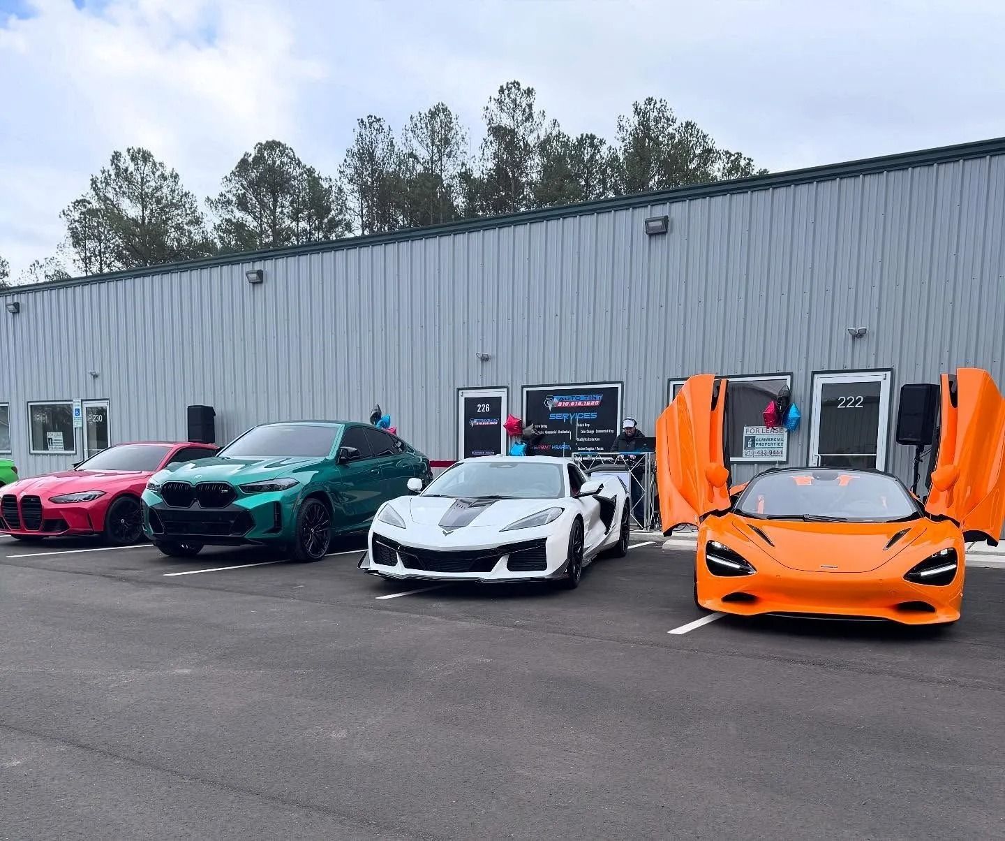 Several colorful luxury cars parked in front of a building: pink, green, white, and orange, one with doors open.