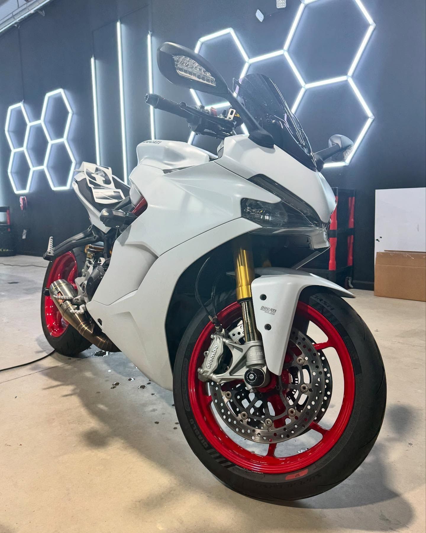 White motorcycle with red wheels, parked indoors, illuminated by hexagonal lights.