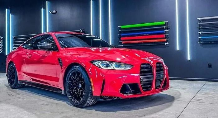 Red BMW M4 coupe parked inside a garage, with colored vinyl rolls on the wall and blue neon lights.