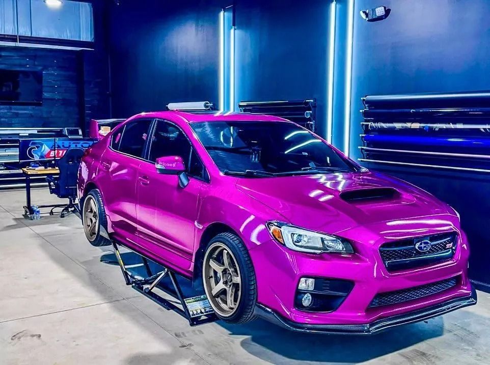Pink Subaru WRX sedan on a car lift in a shop, with rolls of wrap film on the wall.