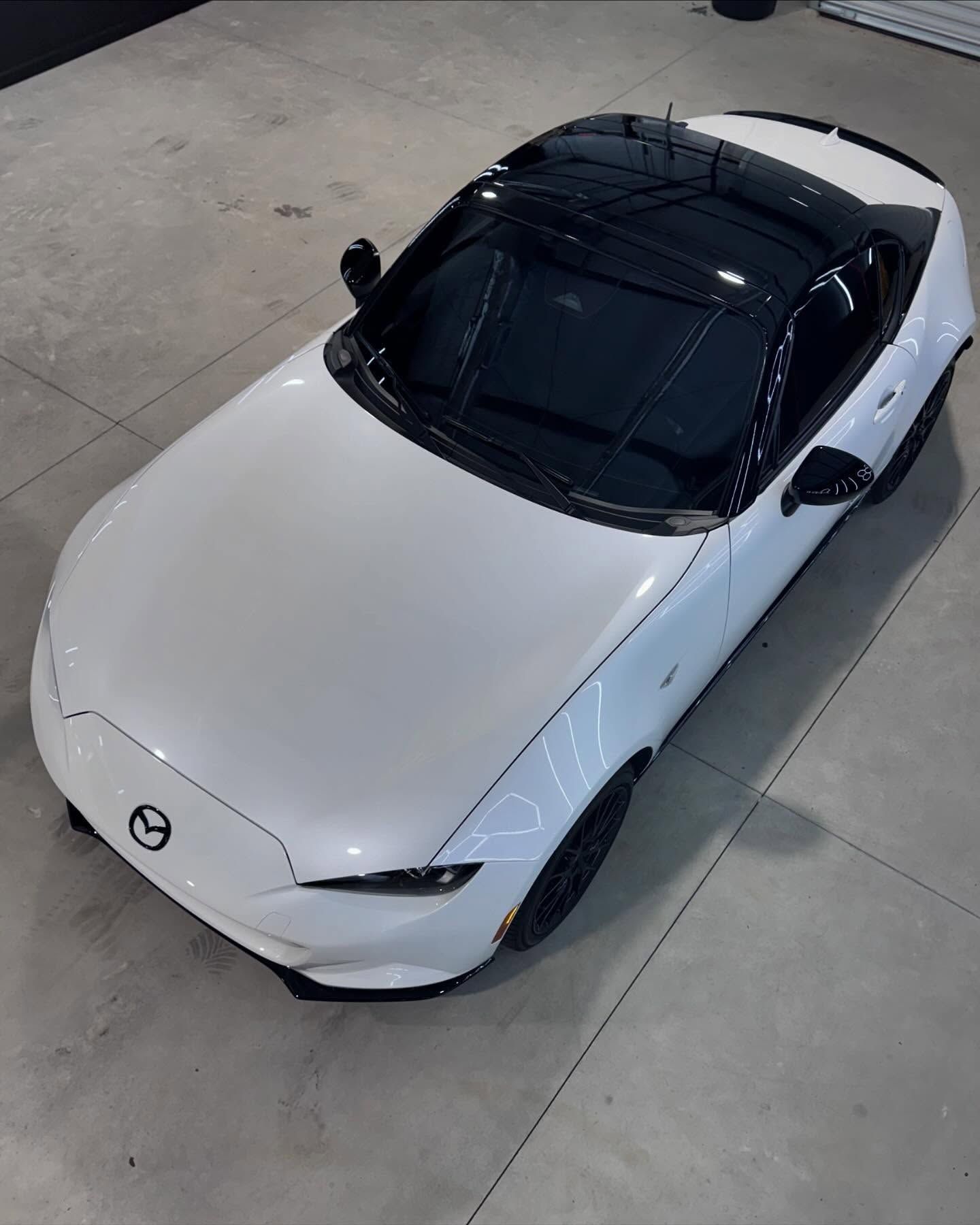 White Mazda MX-5 Miata convertible with black roof and black wheels on a gray surface.