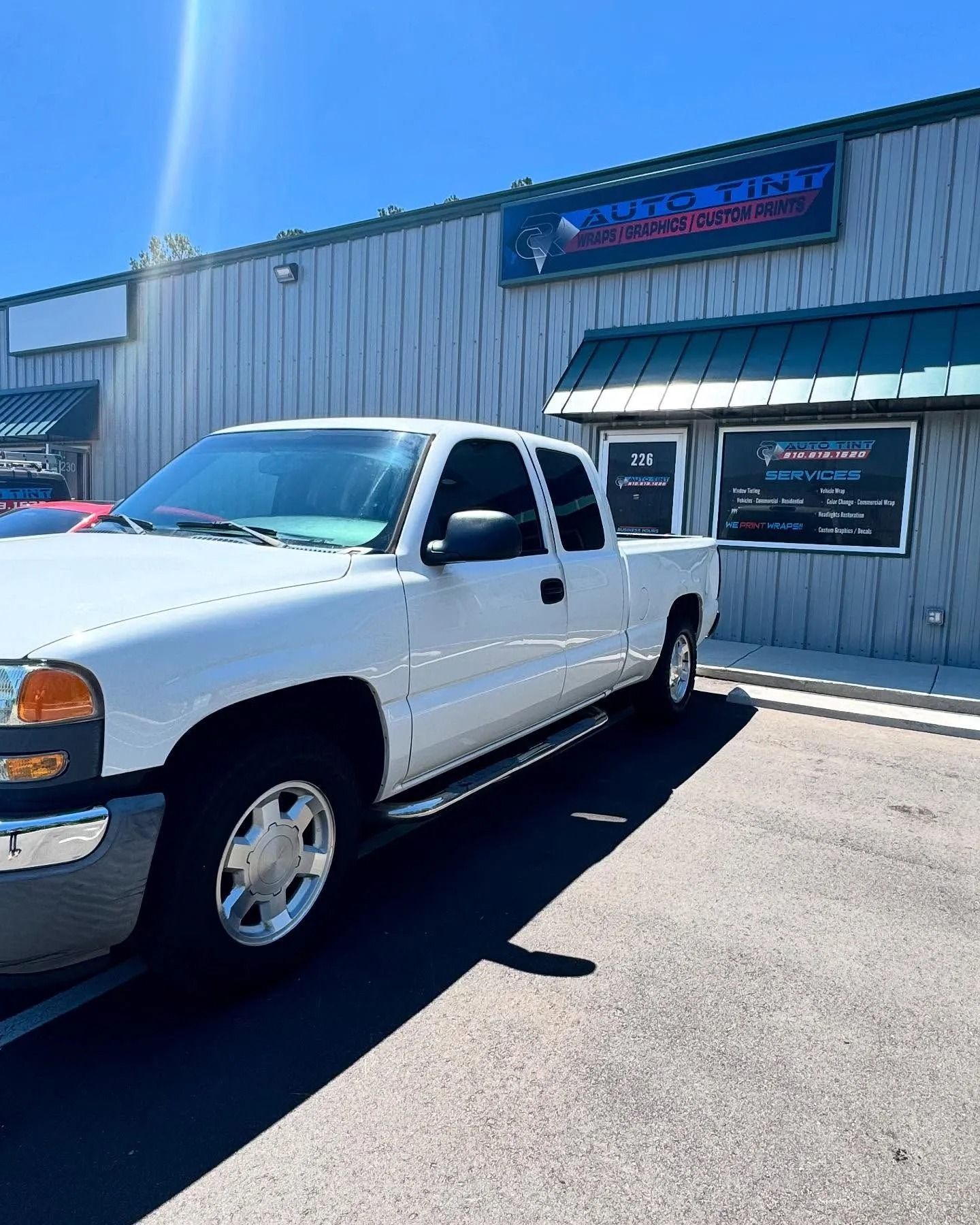 White pickup truck parked in front of an auto shop with blue and green trim and a sign.
