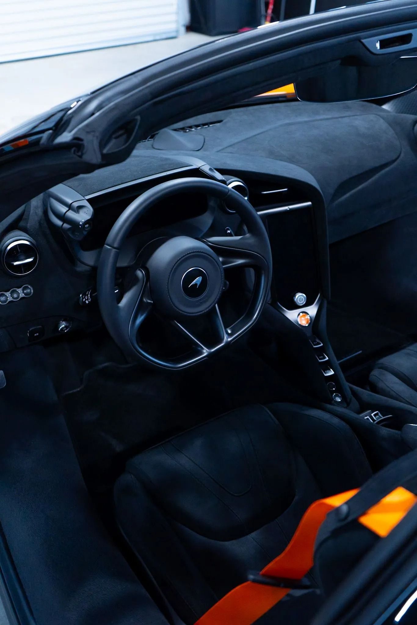 Black McLaren convertible interior with black suede seats, steering wheel, and orange seatbelt.
