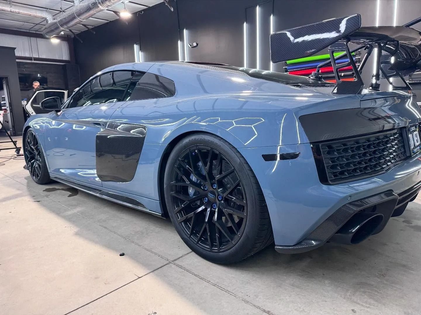 Blue Audi R8 sports car with black rims, spoiler, and carbon fiber accents inside a car shop.