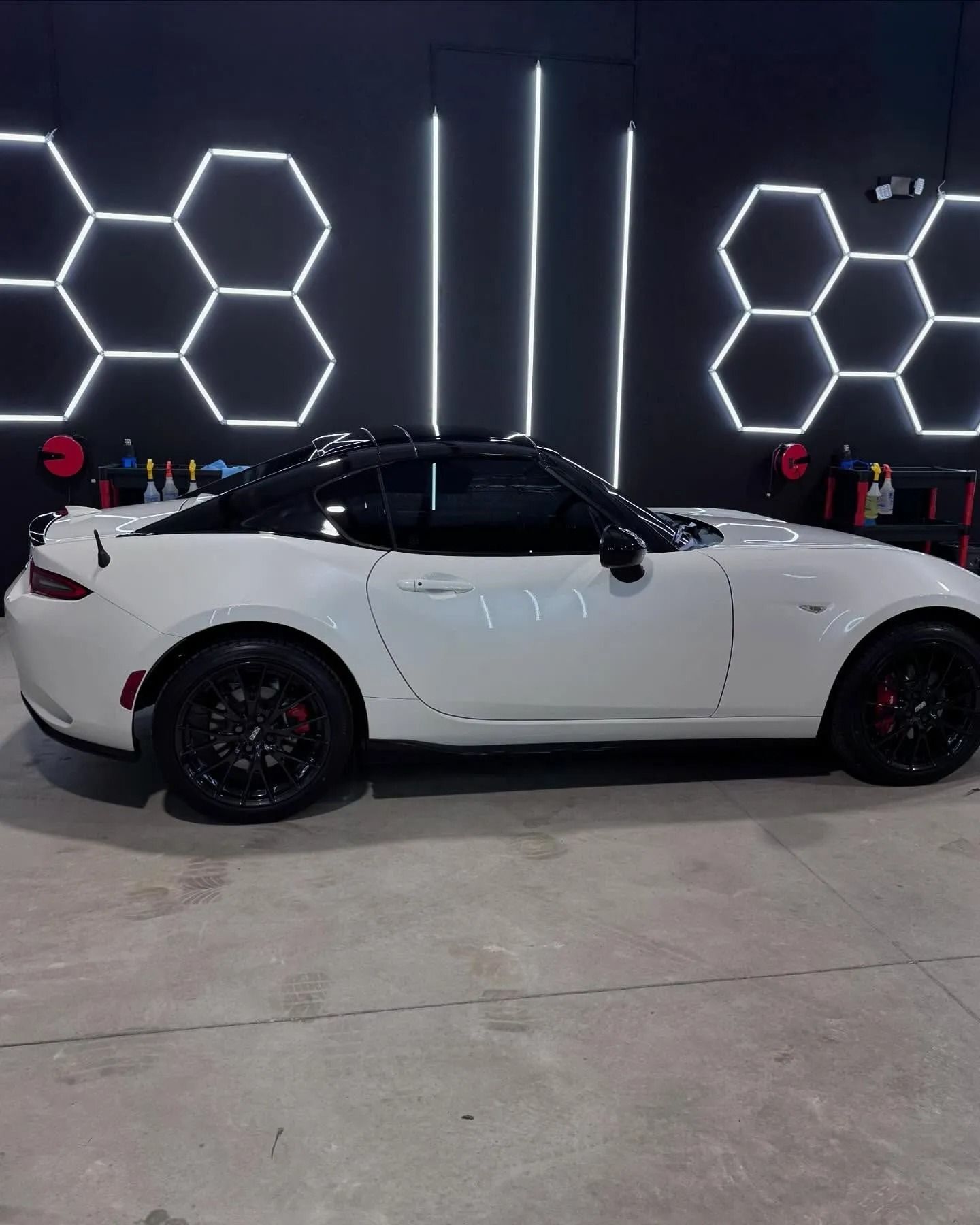 White Mazda Miata sports car with black accents parked in a garage with hexagon and vertical lighting.