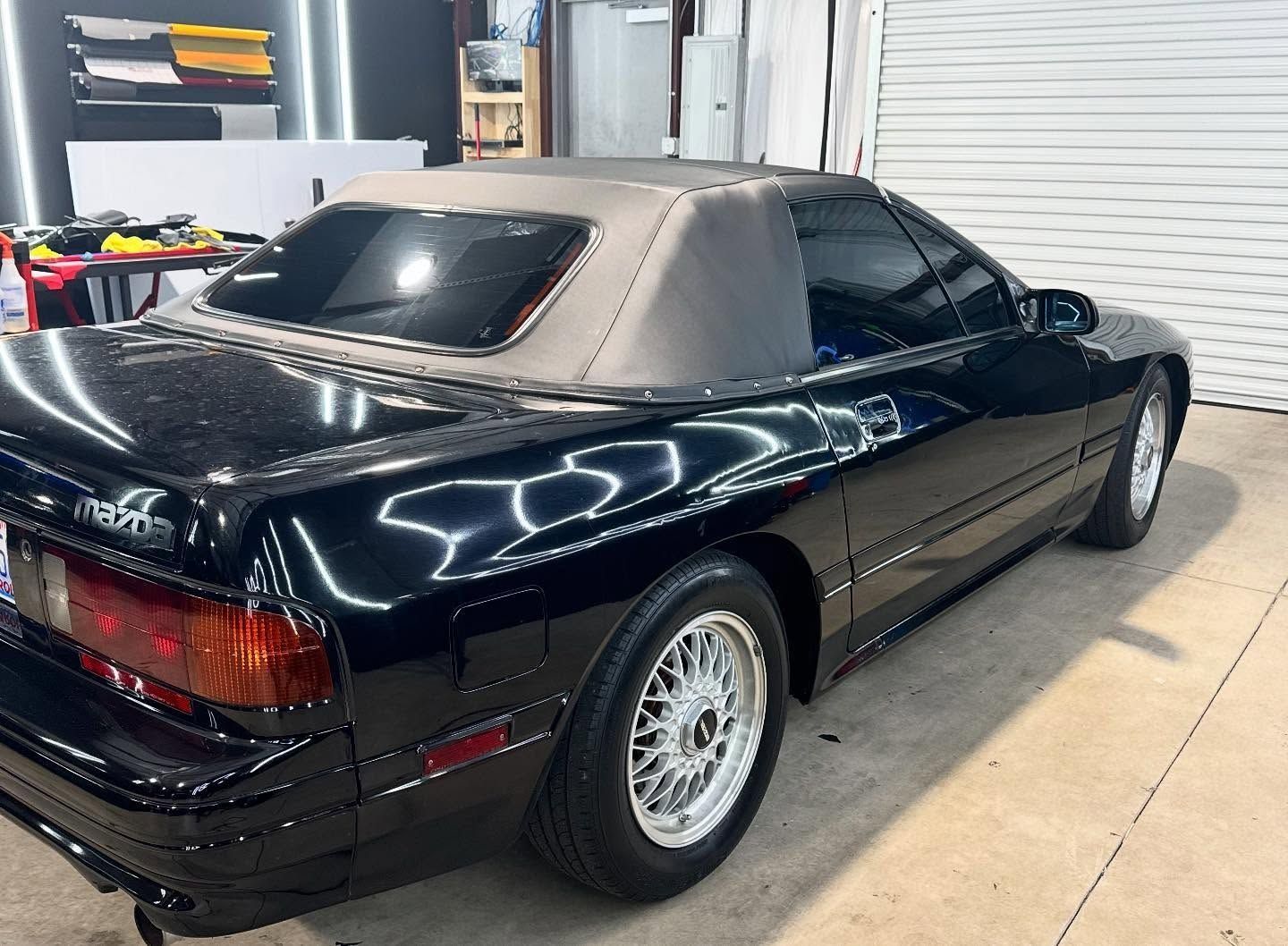 Black Mazda RX-7 convertible parked indoors with a gray soft top and shiny black paint.