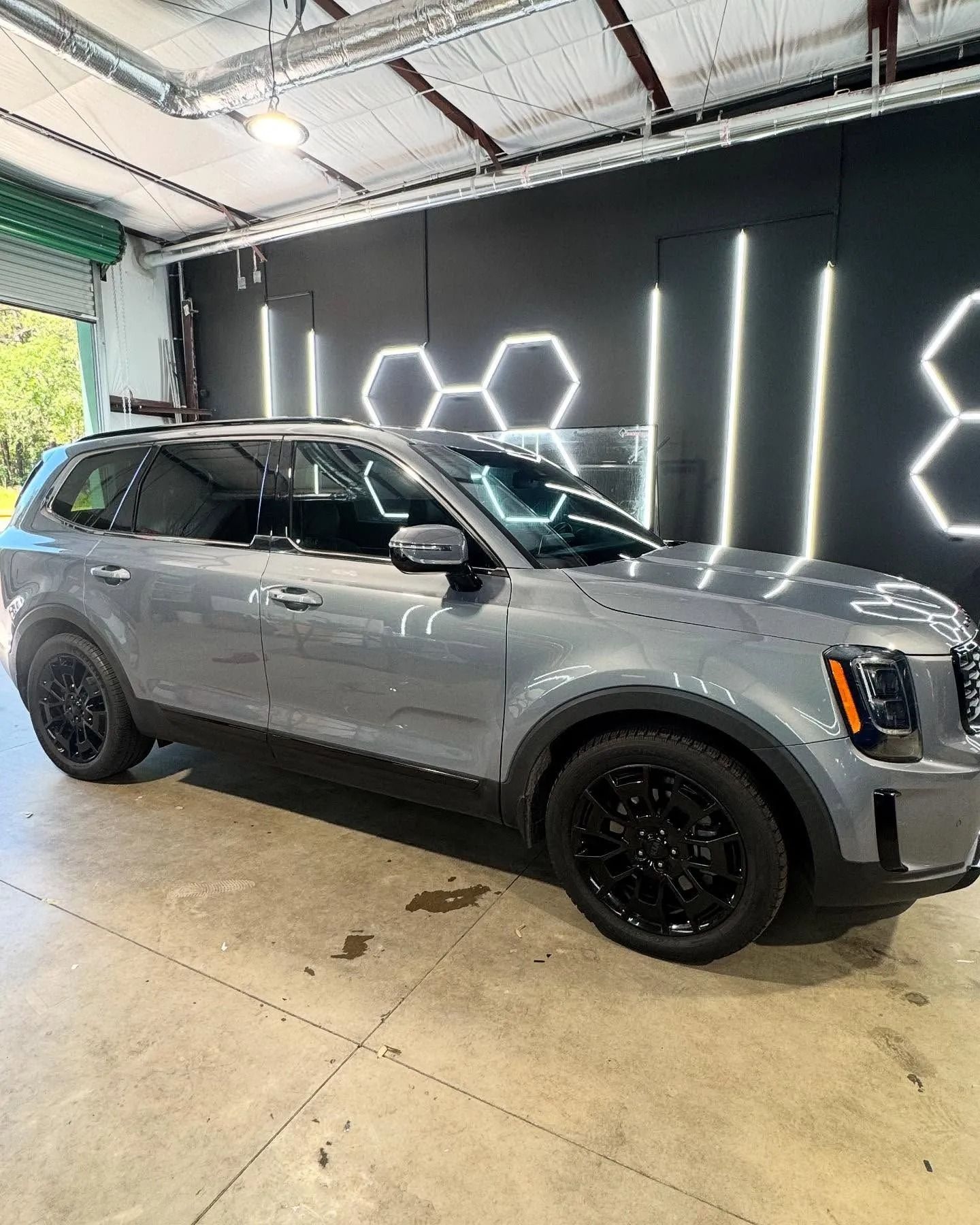 Gray Kia Telluride SUV with black wheels parked inside a garage with black and neon wall decor.