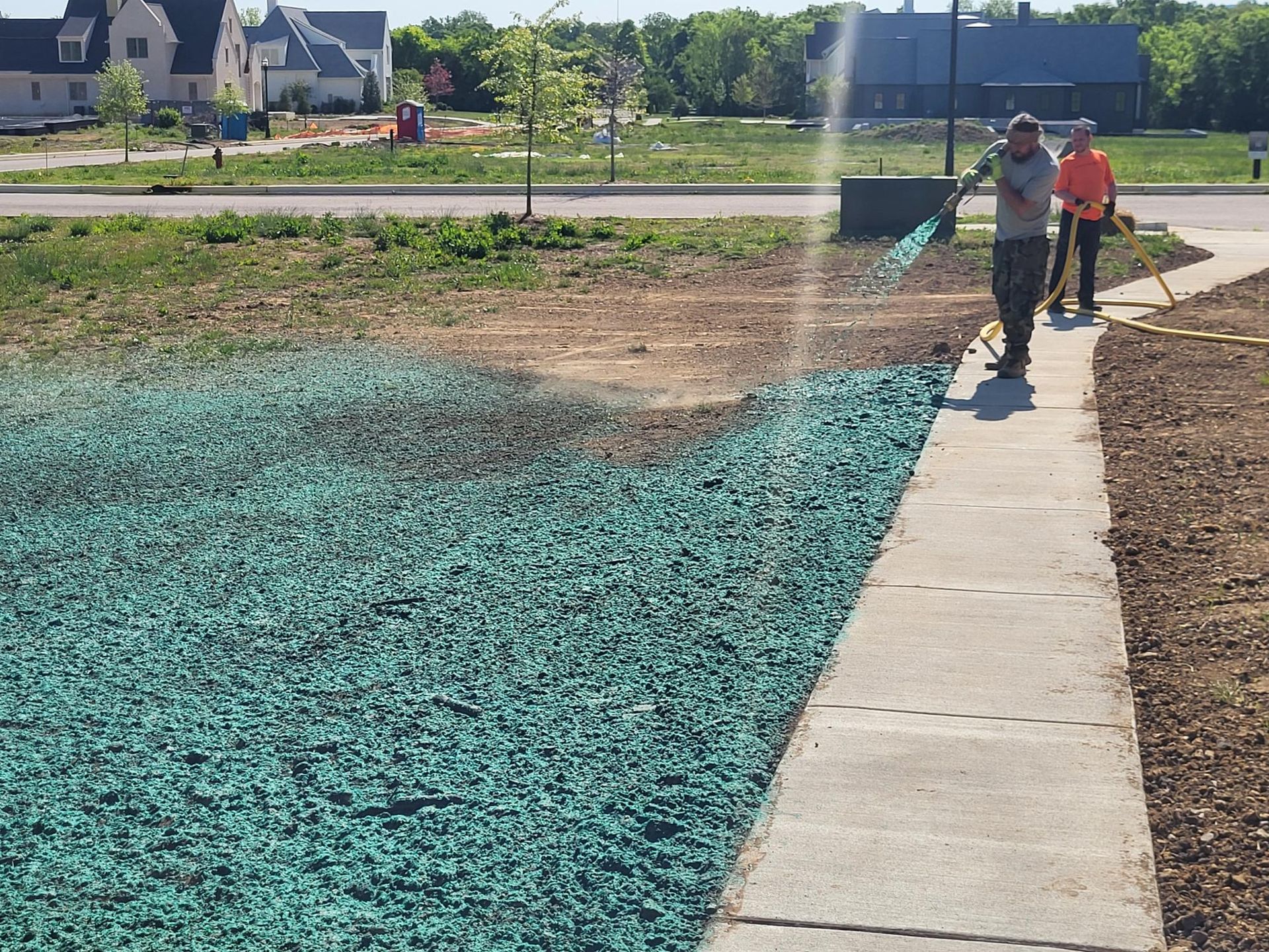 Two workers spraying blue-green coating on a new residential walkway beside a sidewalk