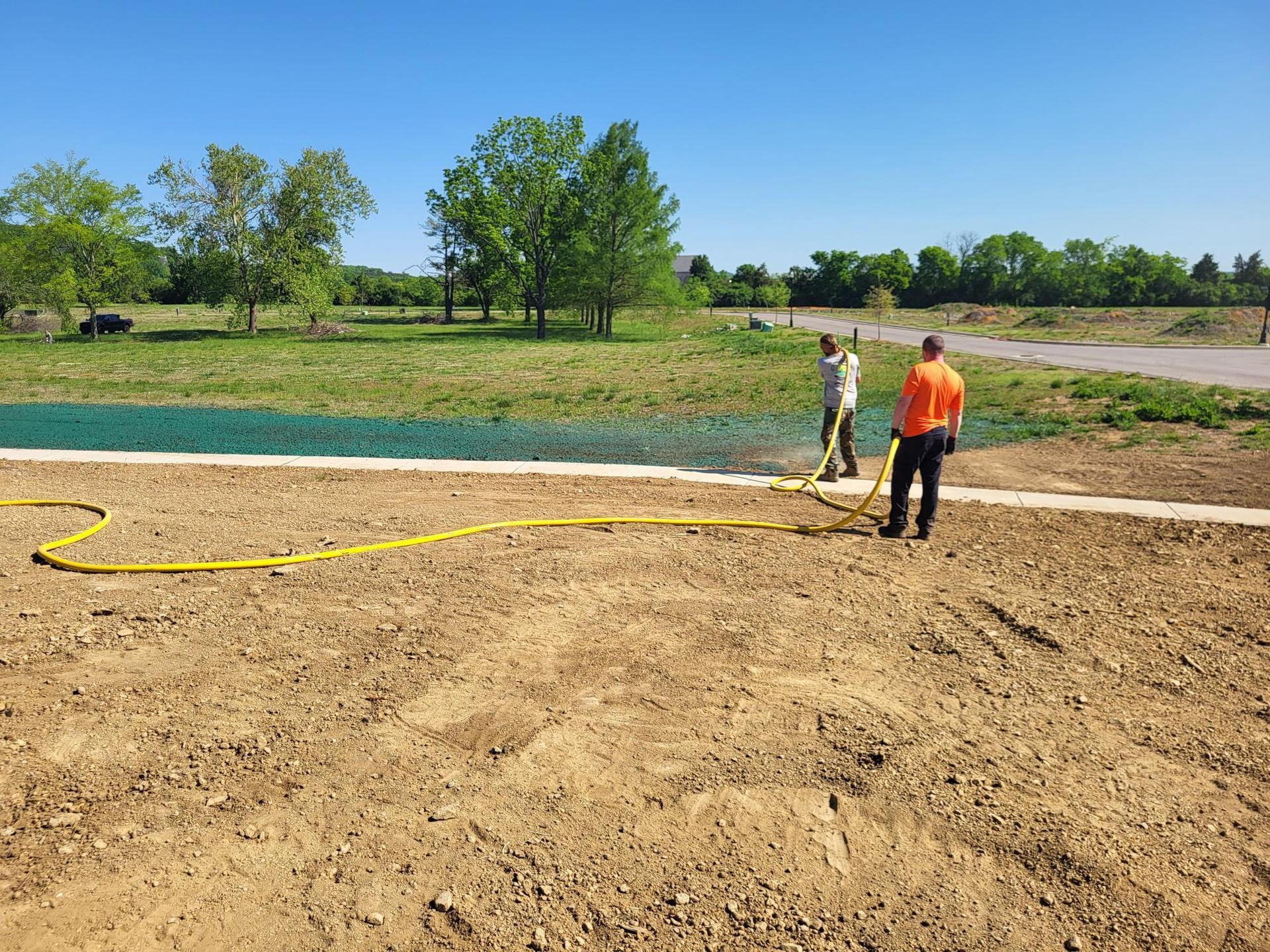 Two workers stand near a hose by a shallow blue-lined pond on a dirt field under clear skies.