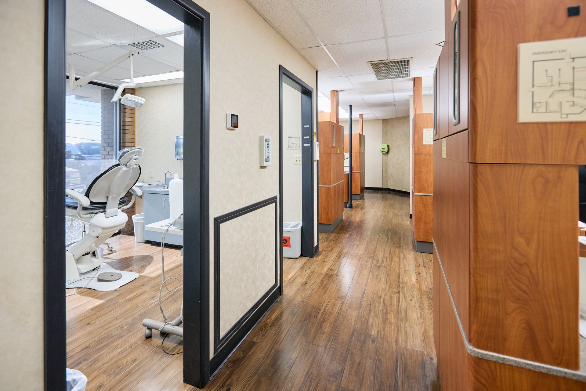 Hallway in a dental office with light wood floors, doors to treatment rooms, and dental equipment visible.