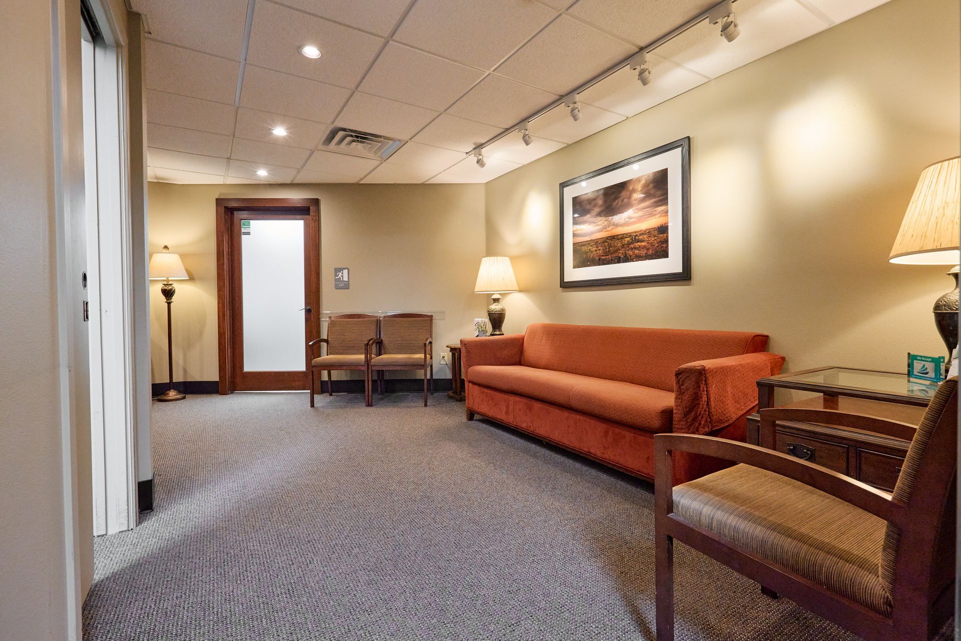 Waiting room with sofa, chairs, artwork, and lamps on beige walls; carpeted floor.