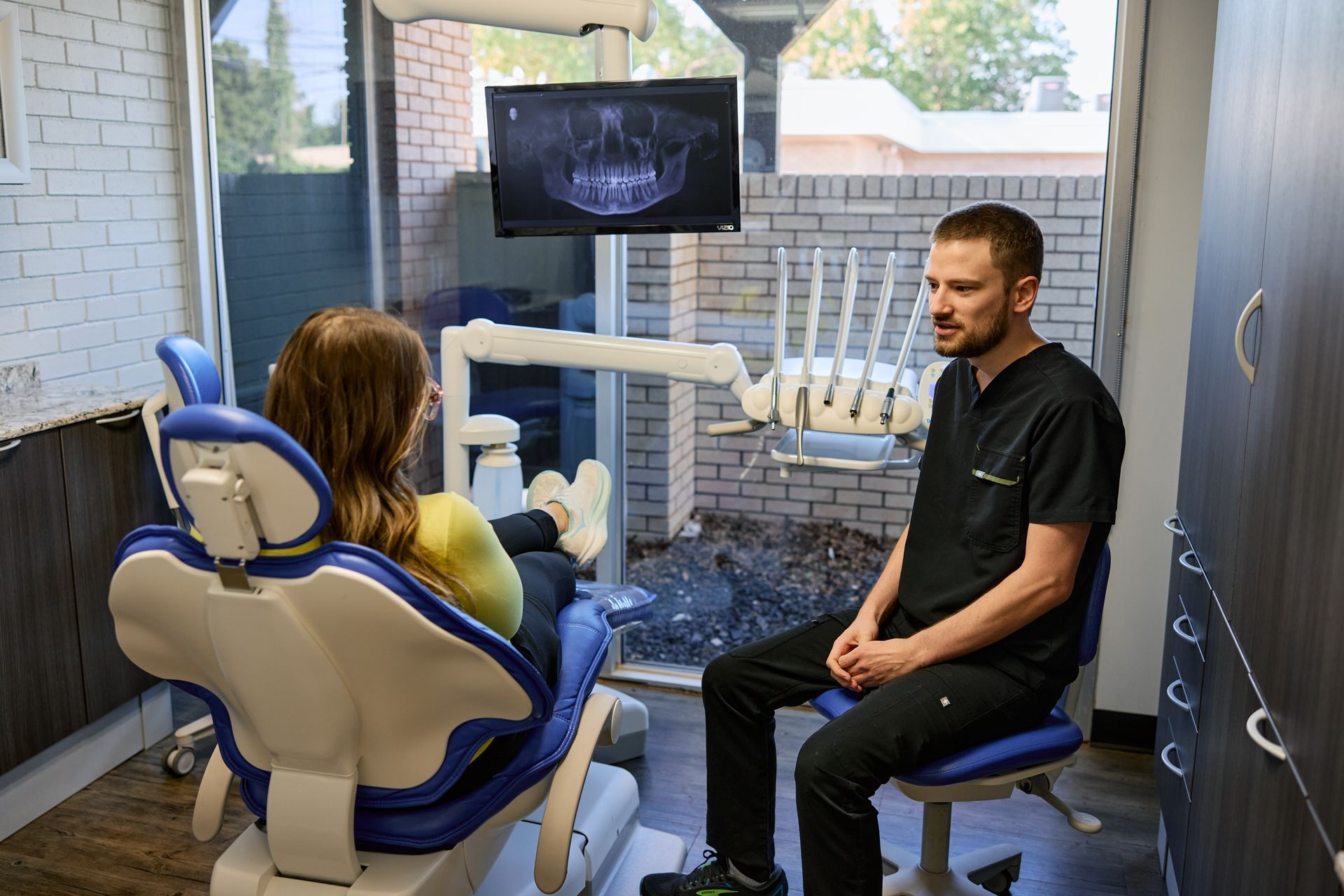 Dentist in black scrubs consults with a patient in a blue dental chair, viewing a dental X-ray.