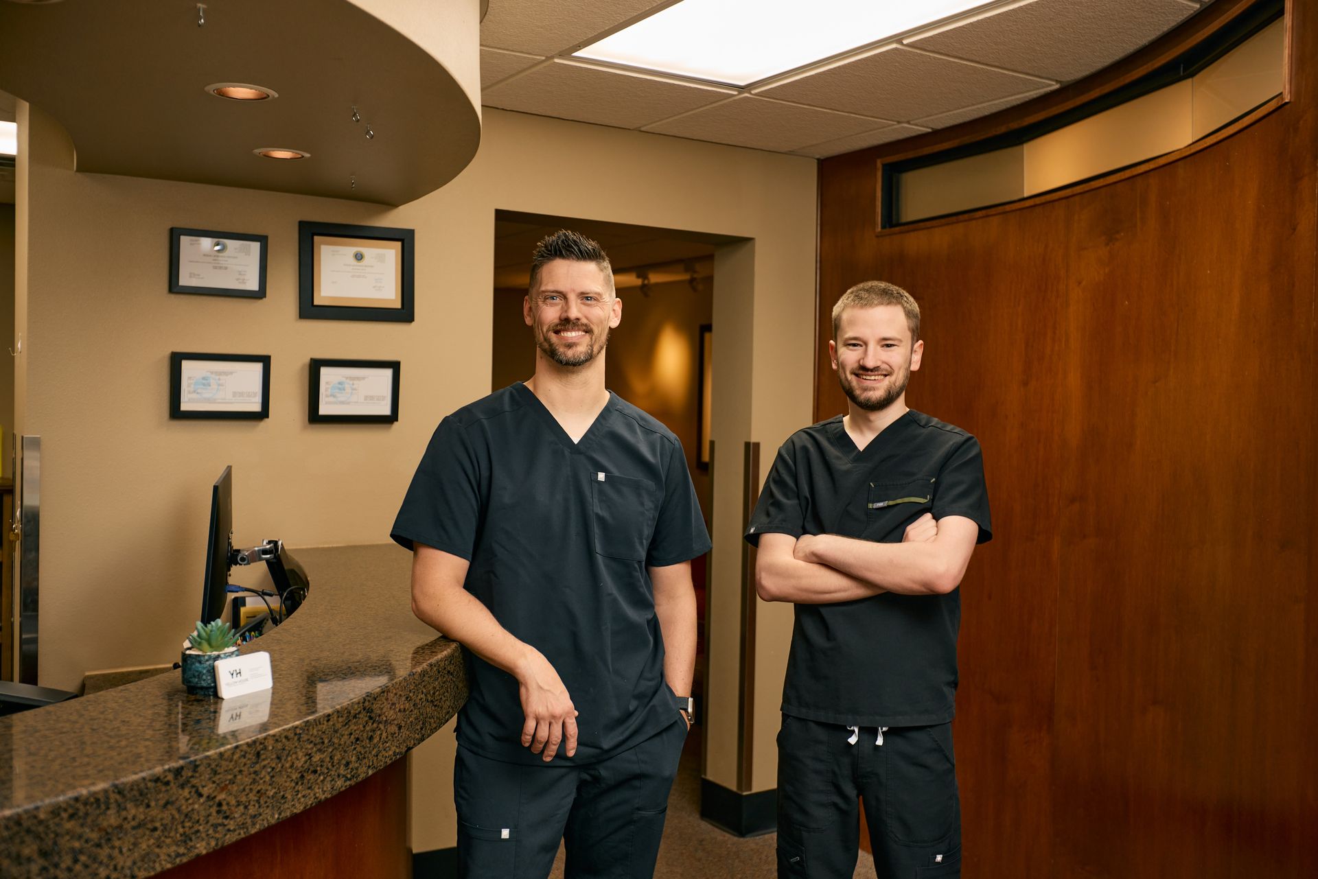 Two men in scrubs standing in an office reception area; one with arms crossed.